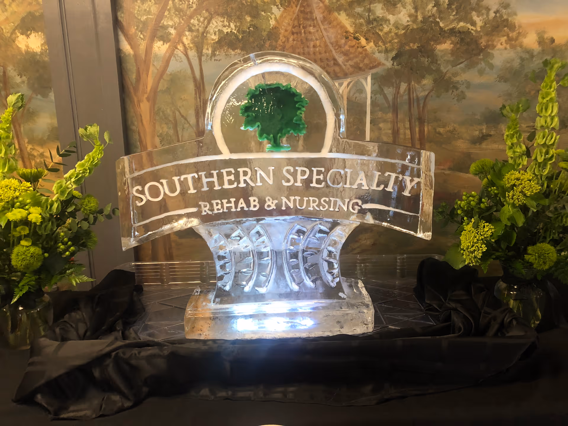An ice sculpture sign reading 'SOUTHERN SPECIALTY REHAB & NURSING' displayed on a table with green floral arrangements and a painted backdrop.
