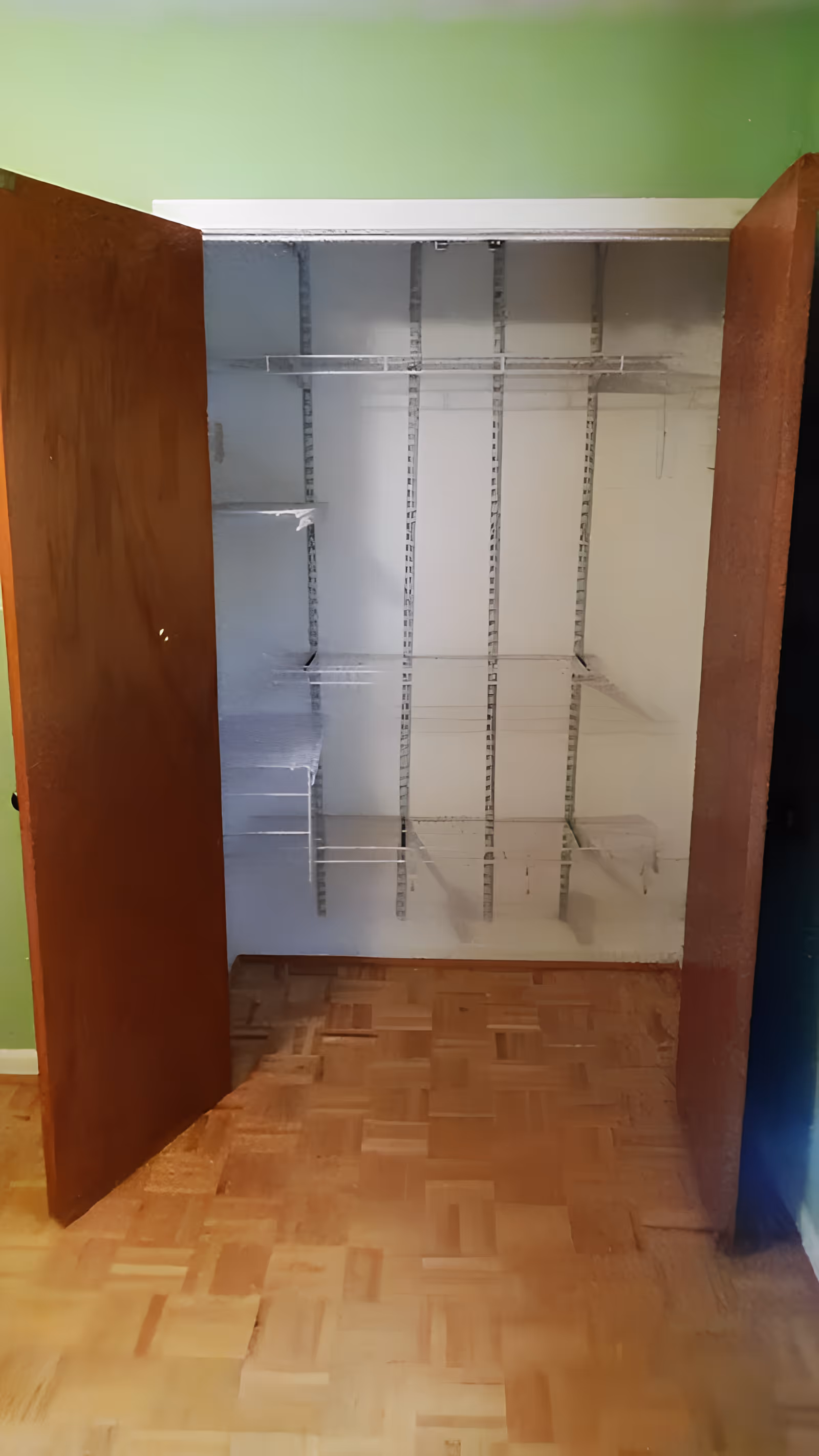 Empty closet with open wooden doors, white interior walls, and metal shelving brackets inside. The closet is set against a green wall and has a parquet wooden floor in front of it.