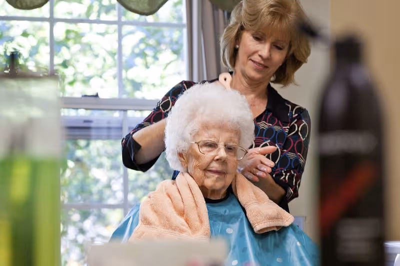 An elderly woman with white hair and glasses is seated wearing a blue cape and a peach towel around her shoulders while a woman standing behind her styles her hair in a bright room with a large window.