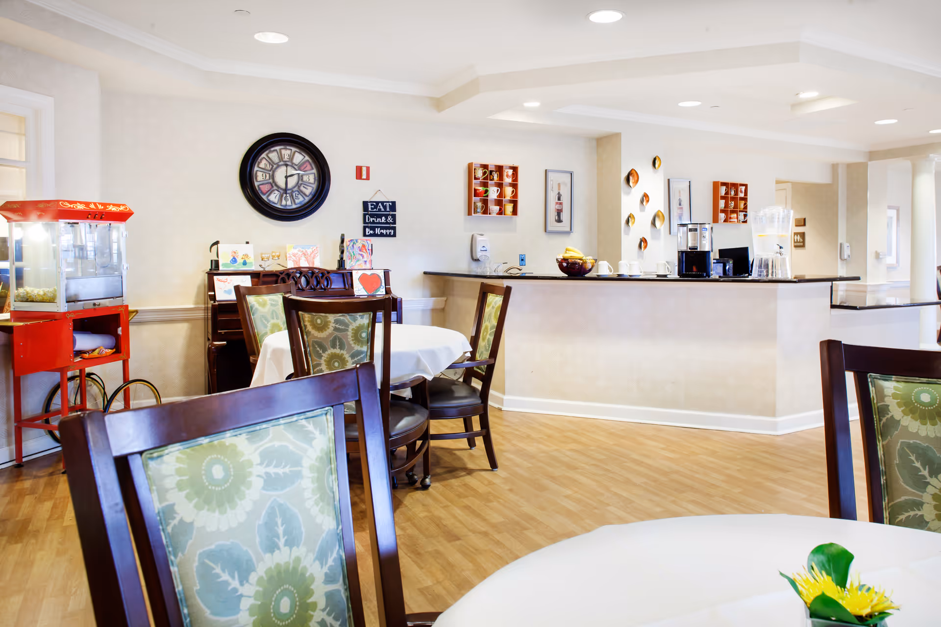 A bright dining area in a senior living facility with round tables covered in white tablecloths and wooden chairs with floral patterned cushions. A red popcorn machine is visible on the left side, and a counter with coffee cups, a coffee machine, and a water dispenser is on the right. The walls are decorated with a large clock, framed pictures, and small shelves holding colorful mugs. The floor is wooden, and the space is well-lit with recessed ceiling lights.