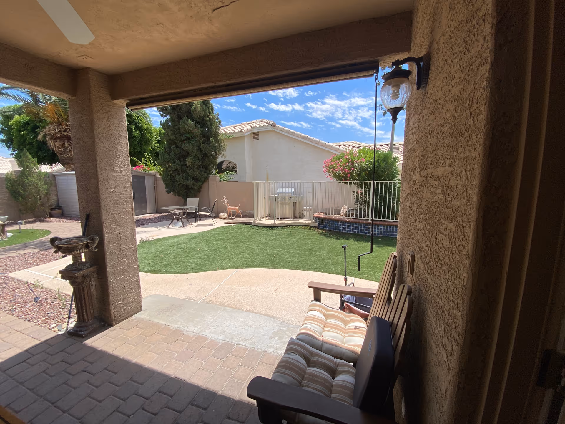 Covered patio overlooking a fenced backyard with artificial turf, patio furniture, and neighboring houses under a blue sky.