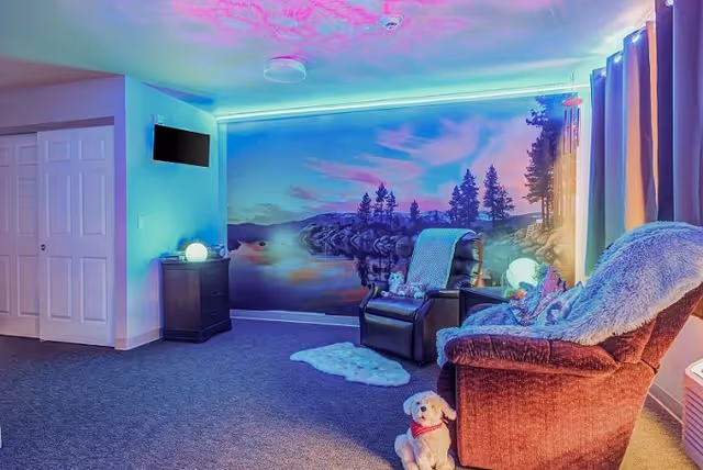 A cozy living room area with two comfortable chairs, one brown and one black, both with blankets draped over them. A small white dog plush toy sits on the carpet in front of the chairs. The wall features a large mural of a serene lake scene with trees and a colorful sky. Soft ambient lighting with blue and purple hues illuminates the room. A small cabinet with a glowing orb lamp and a wall-mounted TV are visible on the left side. Curtains cover a window on the right side.