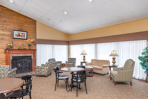 A cozy living room area with multiple seating options including floral armchairs, a beige sofa, and a striped armchair. There are round tables with black chairs, two table lamps on side tables, a fireplace with a wooden mantel, and large windows with sheer white curtains letting in natural light.