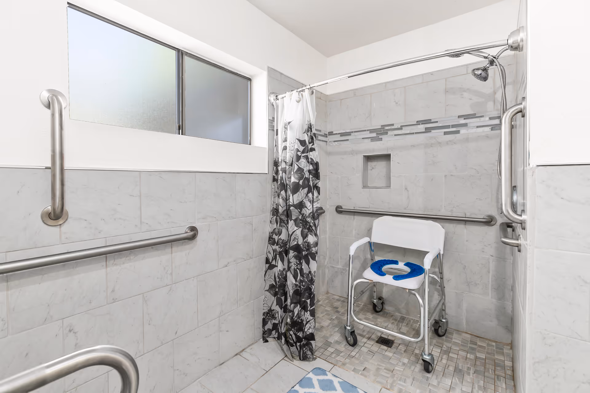 Accessible tiled shower with grab bars, a wheeled shower chair and a patterned shower curtain.