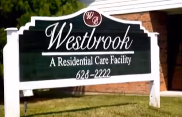 White-framed green outdoor sign in front of a brick building reading "Westbrook A Residential Care Facility 628-2222."