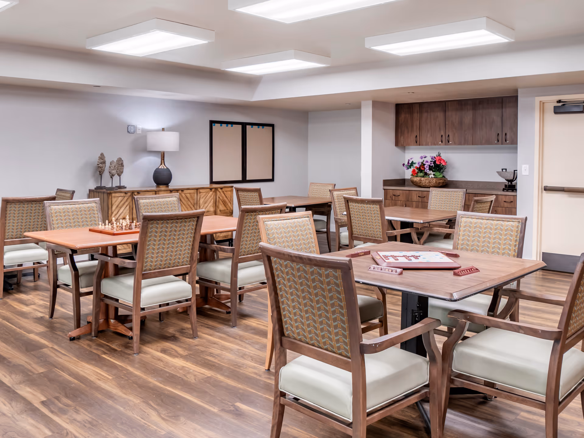 A well-lit activity room with multiple wooden tables and cushioned chairs arranged neatly. One table has a chess set, and another has a Scrabble game set up. The room features wood flooring, a sideboard with a lamp and decorative items, and a cabinet with a basket of flowers. The walls are painted light gray, and there are ceiling lights providing bright illumination.
