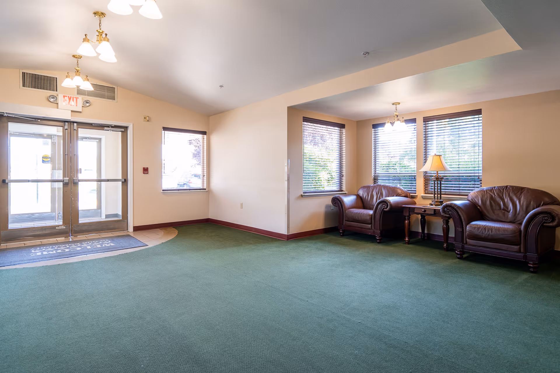A bright lobby area with green carpet, two brown leather armchairs, a wooden side table with a lamp, and large windows with blinds. Double glass doors with an exit sign above are visible on the left side.