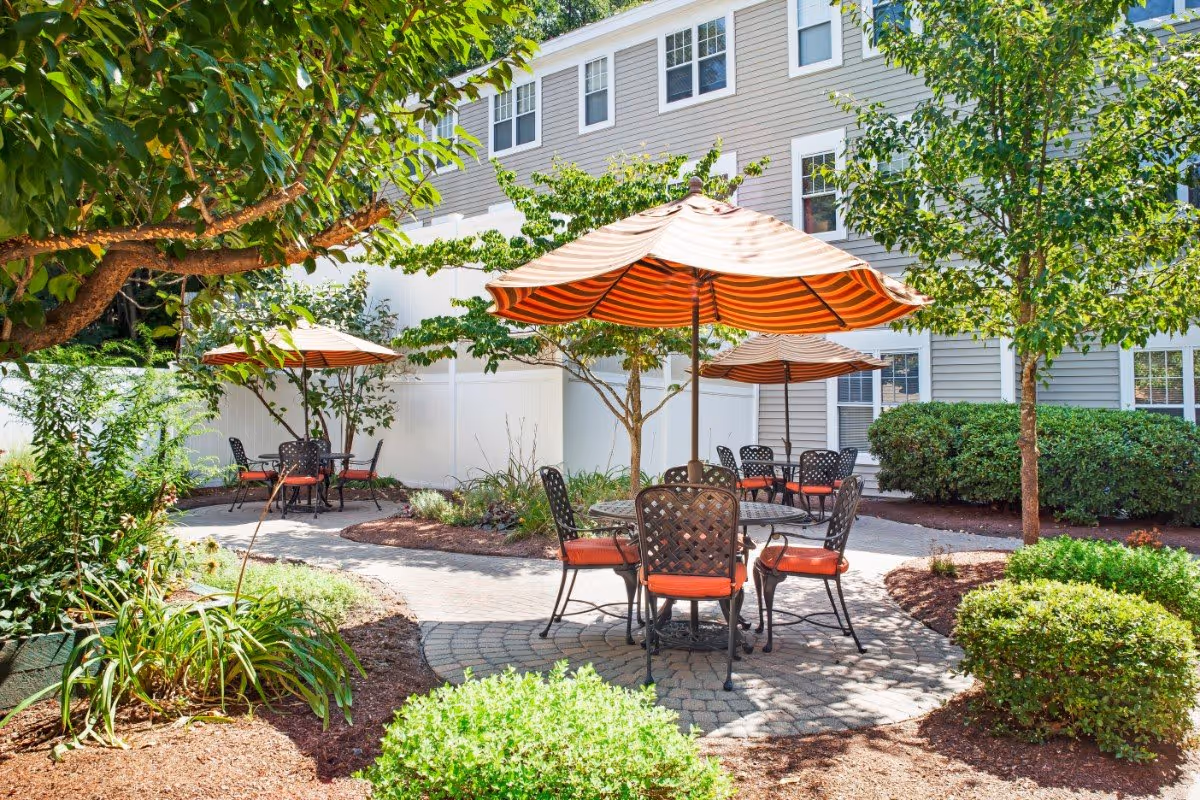 Courtyard patio with round metal tables and orange-striped umbrellas surrounded by trees and shrubs in front of a multi-story building.