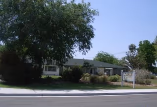 Single-story building with a gray roof surrounded by trees and bushes, with a sign in front that reads 'Lo-Har'. The scene is set on a clear day with a blue sky.