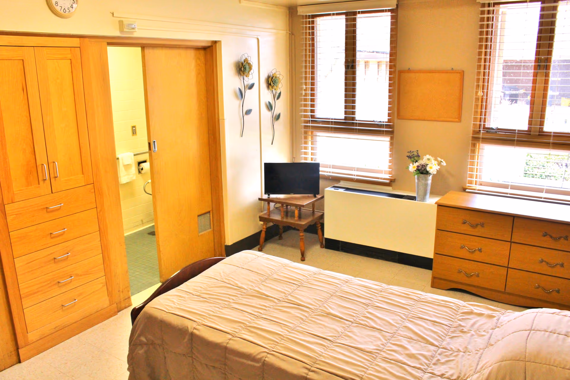 A bright bedroom with a bed covered in a beige quilt, wooden dresser with drawers, a small TV on a wooden stand, and large windows with blinds letting in natural light. There is a wooden sliding door leading to a bathroom with visible towel racks and a toilet paper holder. The room has light-colored walls and a clock above the door.
