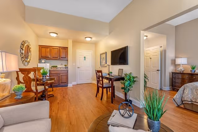 A bright and spacious senior living apartment interior featuring a small kitchen area with wooden cabinets, a dining table with two chairs, a wall-mounted TV, and a cozy living space with a beige armchair and a round coffee table decorated with plants and books. The bedroom is partially visible through an open doorway, showing a bed with a patterned bedspread and a nightstand with a lamp and plant.
