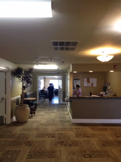 Interior view of a senior care facility hallway with a reception desk on the right where a staff member is working. There is a large potted plant on the left side and a few chairs and tables along the hallway. In the background, a few people are visible in a well-lit room at the end of the hallway.