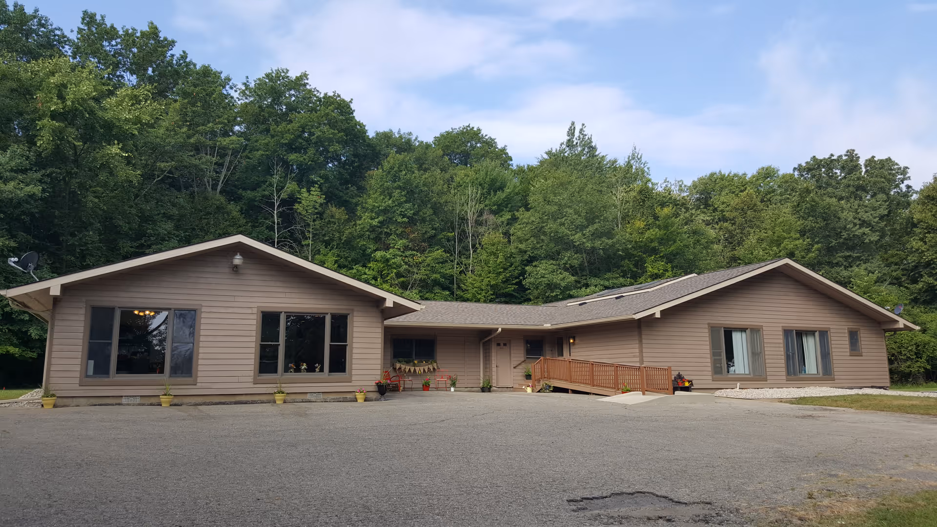 Single-story building with beige siding and a brown shingled roof surrounded by dense green trees. The building has multiple windows and a wooden ramp leading to the entrance. The area in front is paved and spacious.