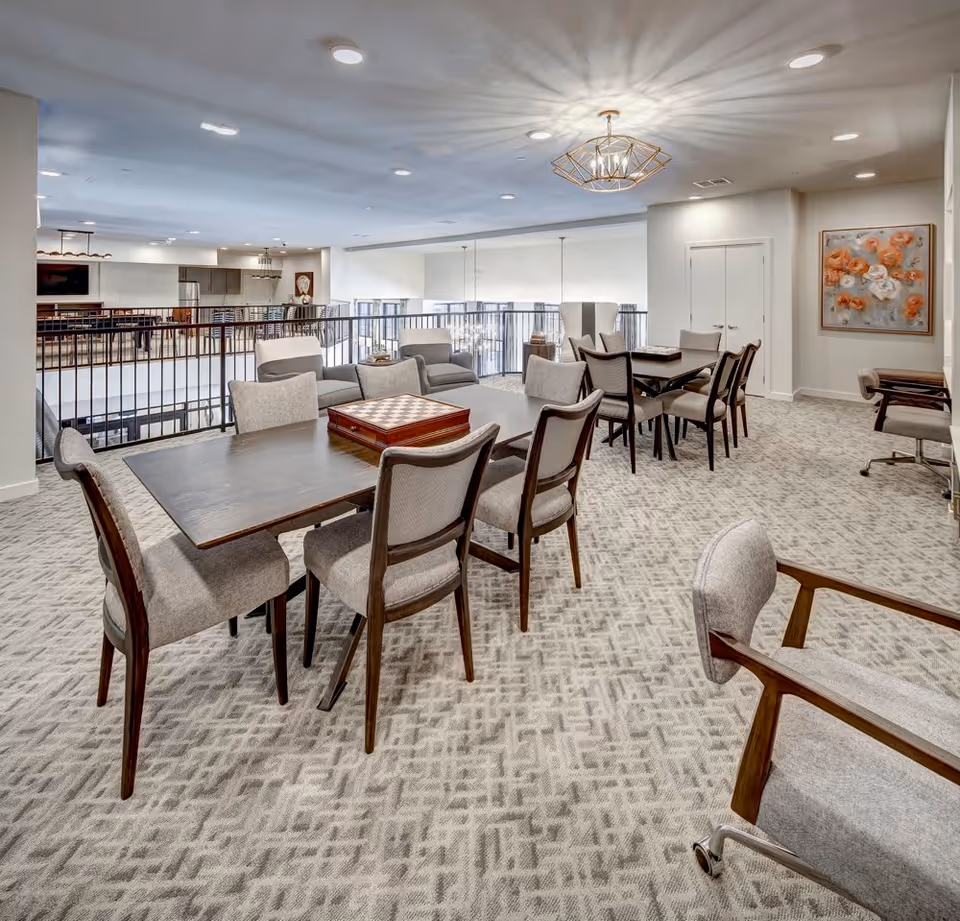 A spacious, well-lit common area with multiple tables and chairs arranged for social or recreational activities. The room features a patterned carpet, modern light fixtures, and a railing overlooking a lower level. There is a painting with orange flowers on the wall and a few armchairs around the tables.