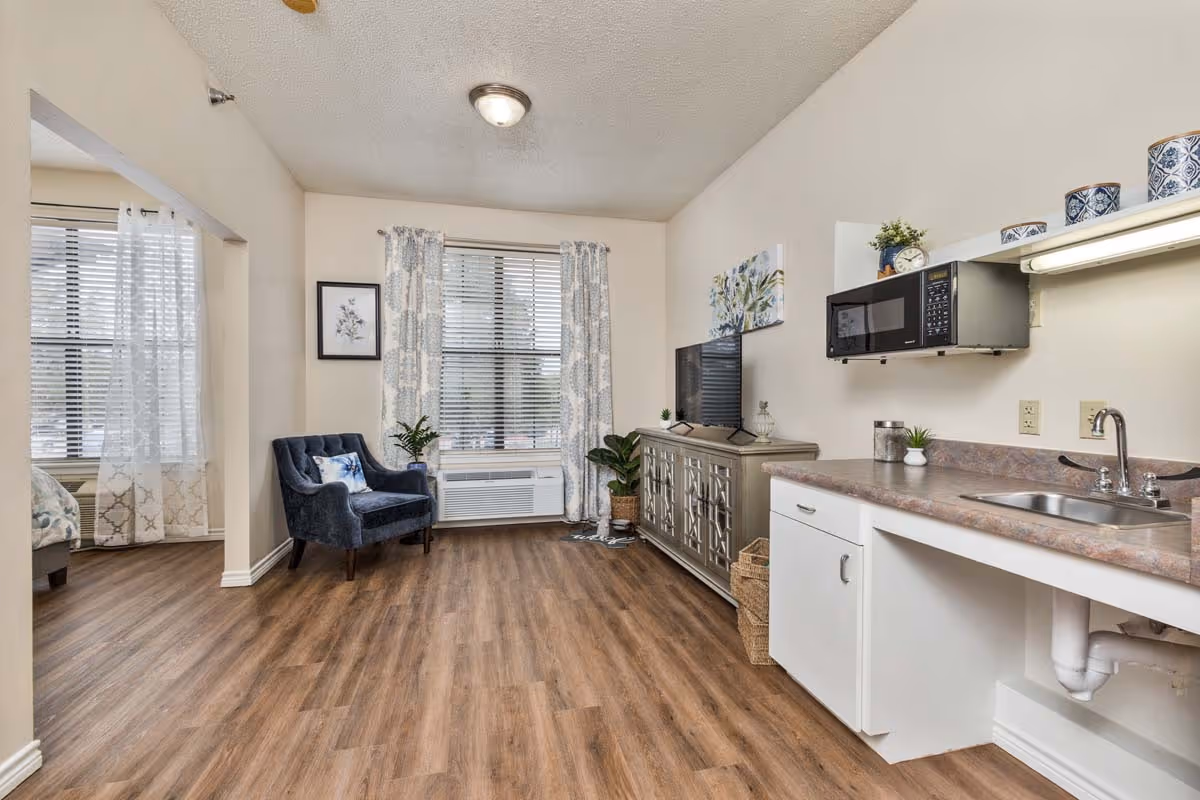 A cozy interior view of a kitchenette area featuring a microwave, sink, and cabinetry, with a comfortable chair and decorative plants nearby.