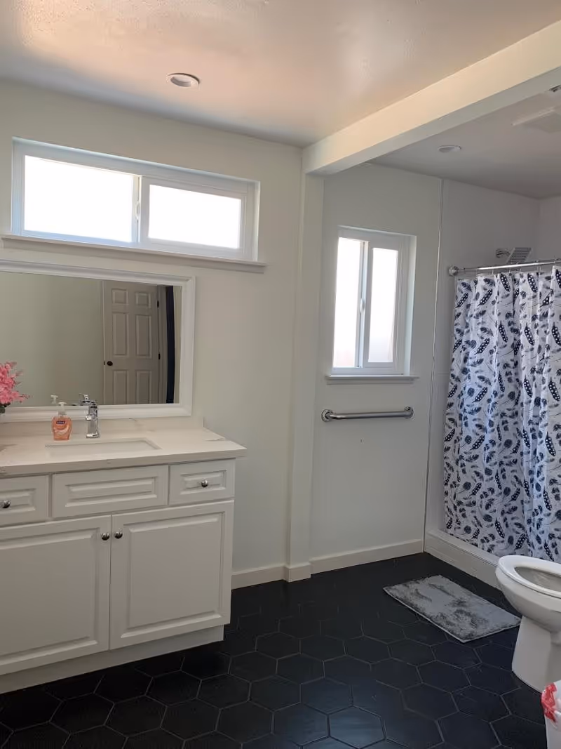 Clean white bathroom featuring a vanity with sink and mirror, a window, a shower with a patterned curtain, a toilet, and a grab bar.