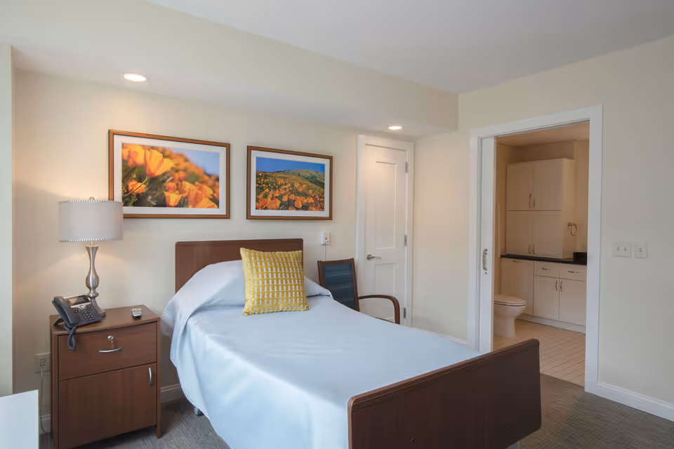 A bedroom in a senior living facility with a single bed covered in a light blue blanket and a yellow patterned pillow. Next to the bed is a wooden nightstand with a lamp, telephone, and remote control. Two framed pictures of orange flowers hang on the wall above the bed. A chair is placed near a closed door, and an open doorway reveals a bathroom with white cabinets and a toilet.