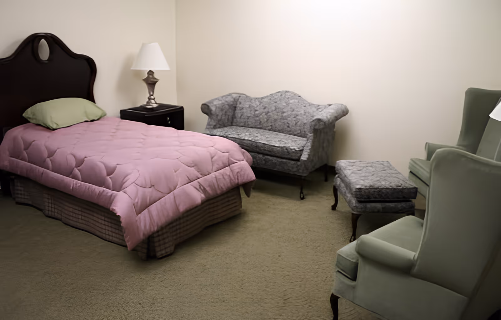 A bedroom with a single bed covered with a pink quilt and a green pillow, a dark wooden headboard, a nightstand with a lamp, a small gray loveseat, a matching ottoman, and two green armchairs on a beige carpeted floor.