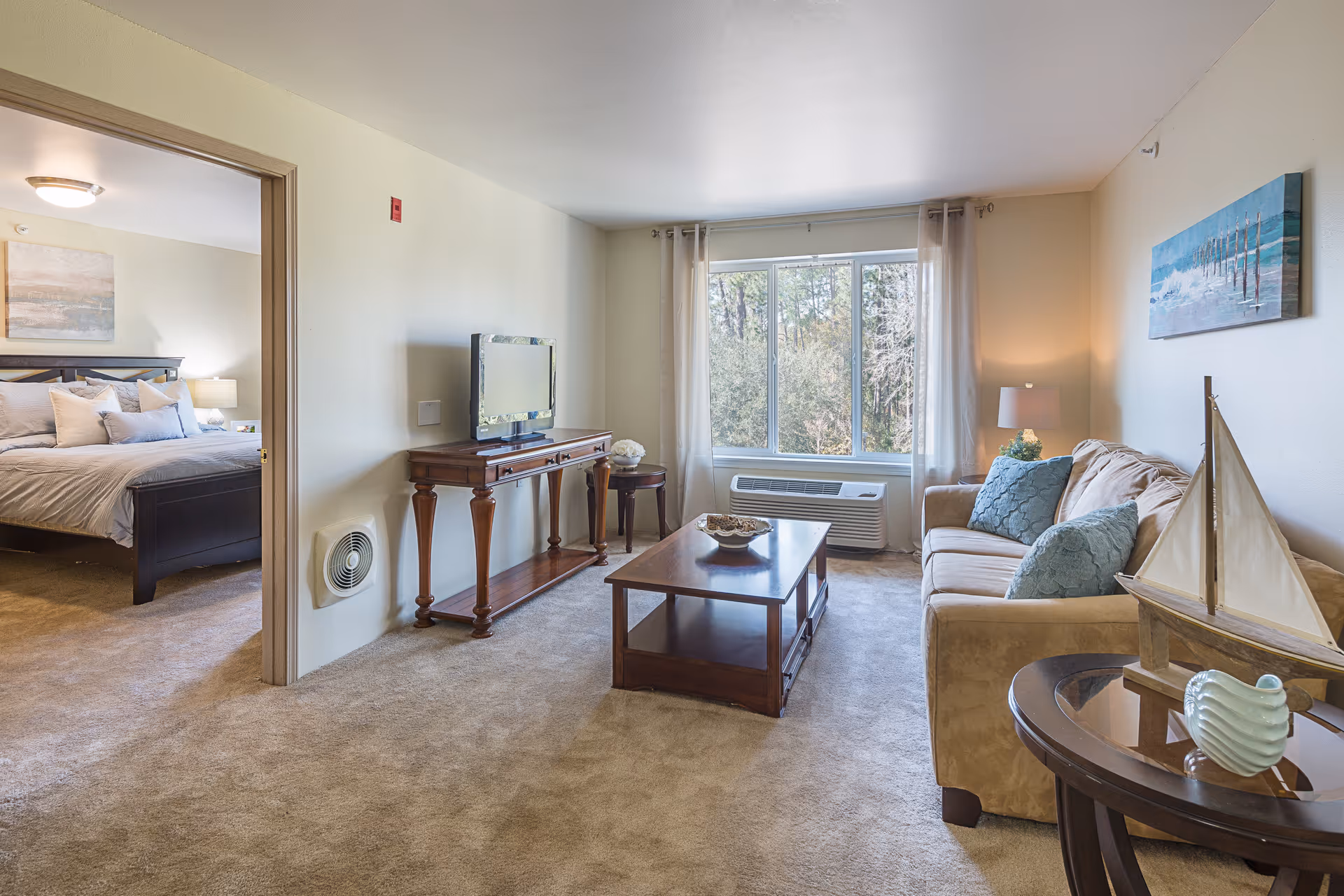 A cozy living room with a beige sofa adorned with blue cushions, a wooden coffee table, and a TV on a wooden console table. A large window with sheer curtains lets in natural light and shows trees outside. To the left, an open doorway leads to a bedroom with a neatly made bed and a bedside lamp. The room has beige carpet and light-colored walls, with a decorative sailboat and a small vase on a round side table.