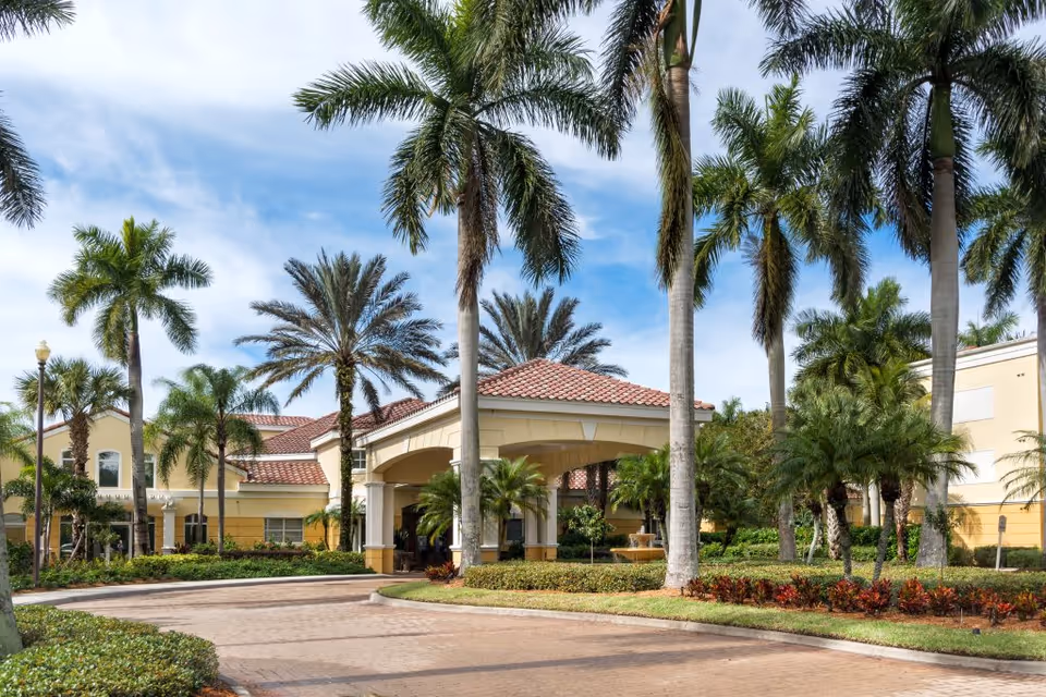 Front entrance of The Carlisle Naples with a covered porte-cochere, palm trees, and a landscaped circular driveway.