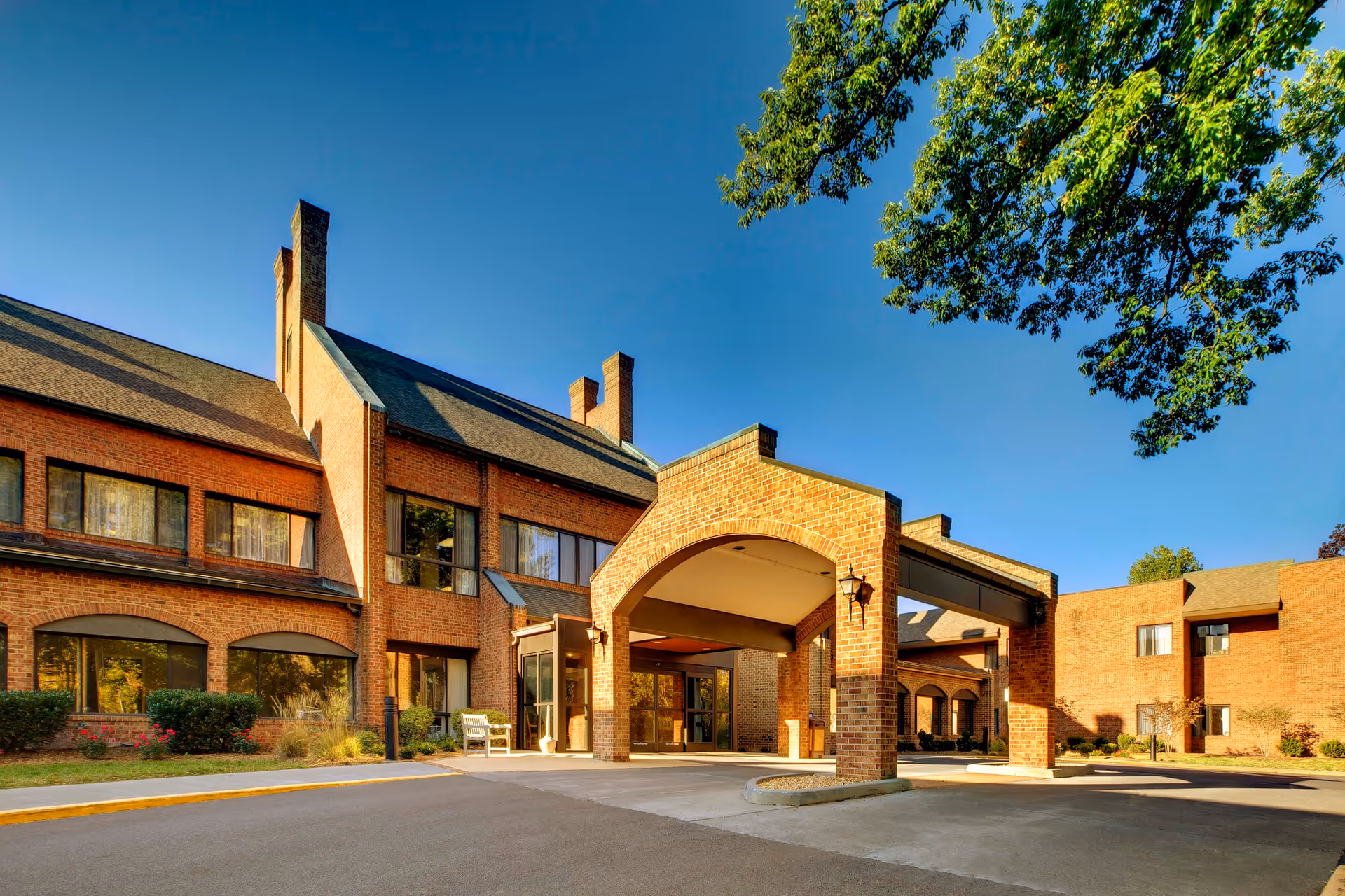 Front entrance of a brick senior living community with a covered porte-cochère under a clear blue sky.