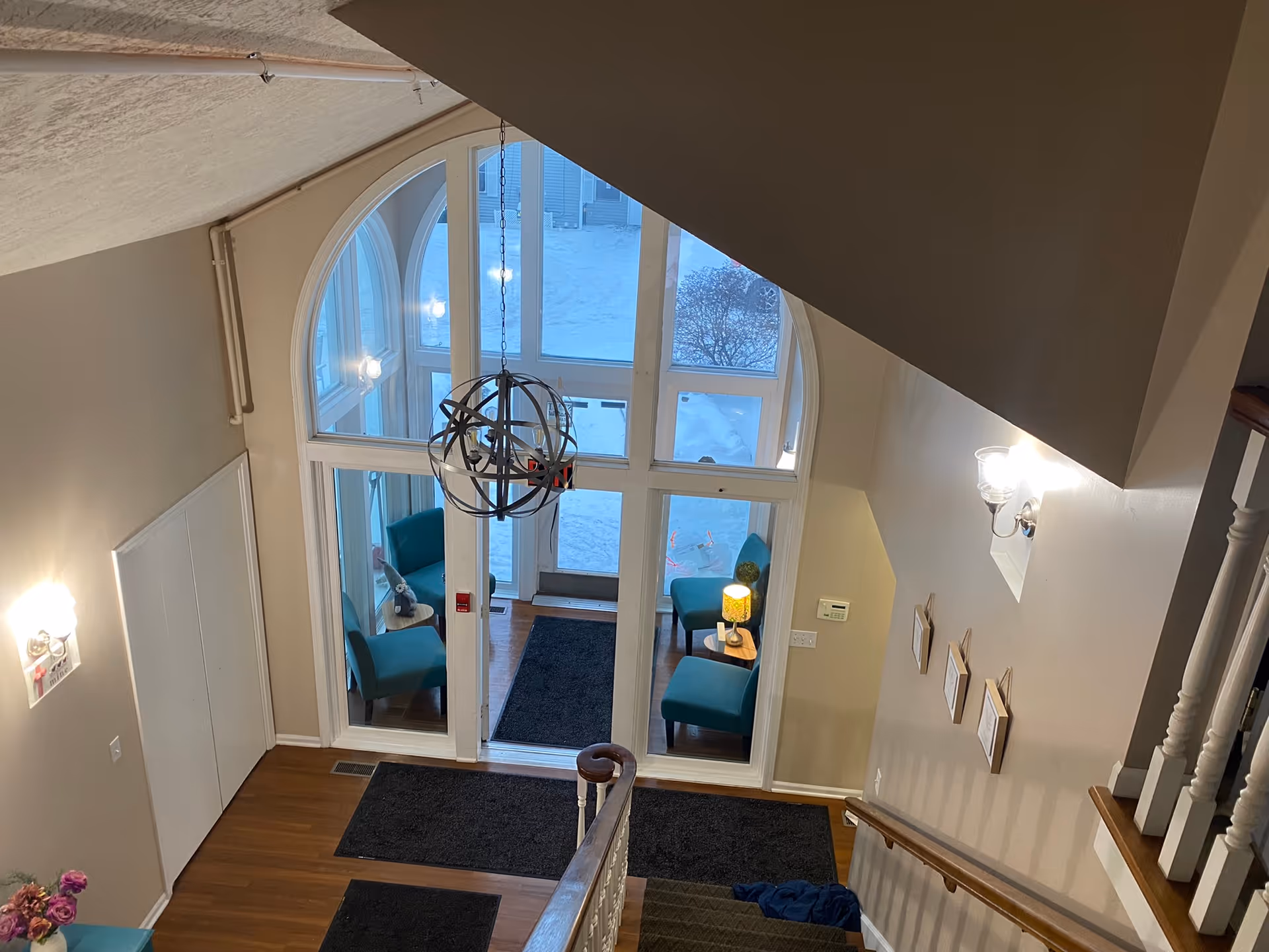 View from the top of a staircase looking down into a bright entryway with large arched windows. The entryway has teal chairs, small tables with lamps, and dark floor mats on a wooden floor. Snow is visible outside through the windows.