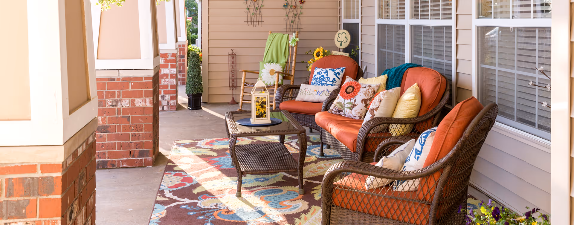 A cozy outdoor patio area with wicker furniture including two chairs and a loveseat with orange cushions and decorative pillows. A wicker coffee table with a lantern sits on a colorful patterned rug. The patio is adjacent to a building with brick and beige siding, and there are potted plants and decorative items around.