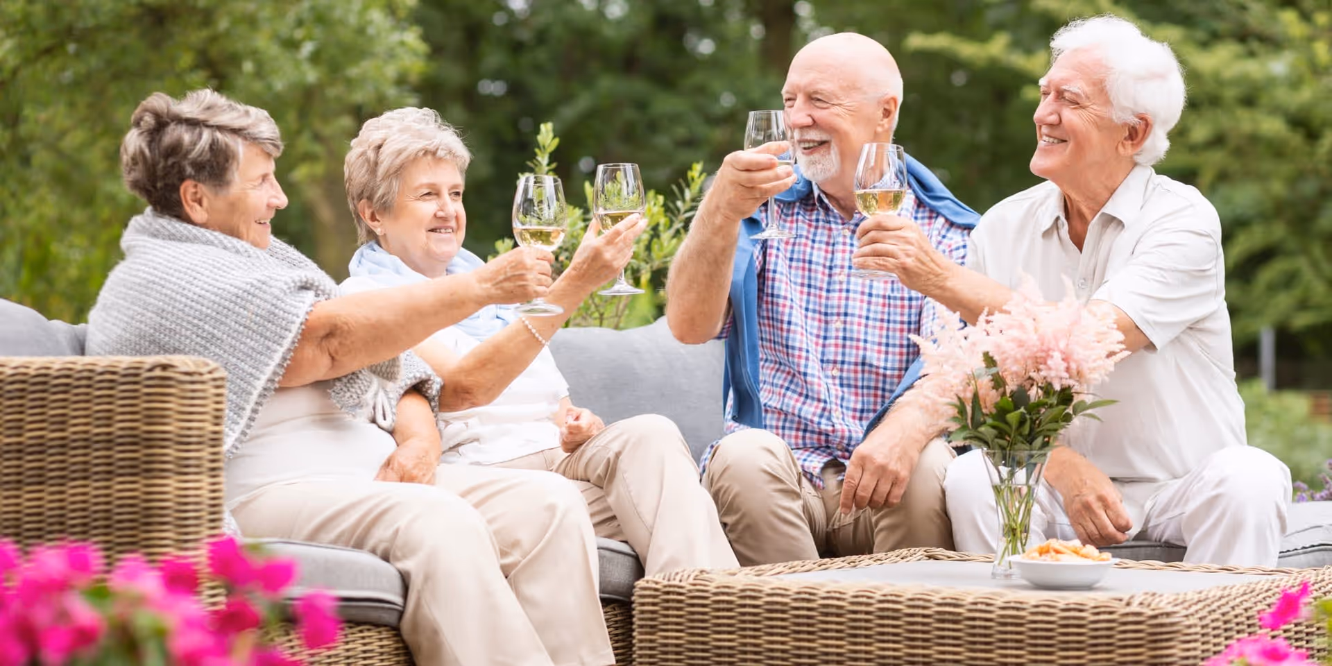 Four elderly people sitting outdoors on a wicker sofa and chairs, raising glasses of white wine in a toast, with a table in front of them holding a vase of flowers and a bowl of snacks, surrounded by greenery and pink flowers.