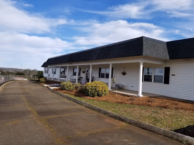Exterior view of a single-story building with white siding and a black roof, featuring a covered porch with white columns and several windows. There are bushes and a small garden area along the porch, and a paved driveway or walkway runs alongside the building under a partly cloudy sky.
