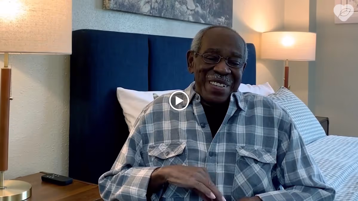 An elderly man wearing glasses and a plaid shirt is sitting on a bed with a blue upholstered headboard. There are two bedside lamps on either side of the bed, and pillows with checkered pillowcases are visible behind him. The room has a calm and cozy atmosphere.