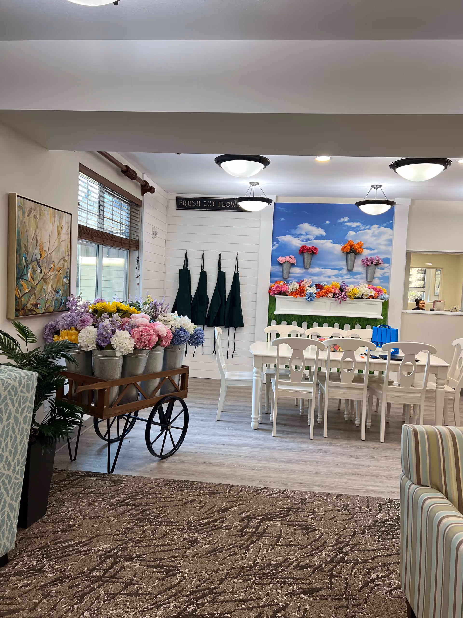 Interior room with a wooden cart holding metal buckets filled with colorful artificial flowers. A white table with white chairs is set against a wall decorated with a mural of a blue sky and clouds, with flower pots attached. Four dark green aprons hang on the wall under a sign that reads 'FRESH CUT FLOWERS'. The room has light wood flooring, a patterned carpet, and ceiling lights.