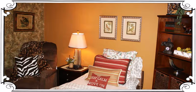A cozy bedroom corner with a single bed adorned with multiple pillows, including one with the text 'KEEP CALM & MOVE ON'. Next to the bed is a dark armchair with a zebra-patterned pillow and a leopard print throw. A small nightstand with a lamp and a small plant sits between the bed and chair. The walls are painted warm yellow and decorated with two framed bird pictures. A wooden bookshelf with decorative items and books is visible on the right side. The image has a decorative white border.