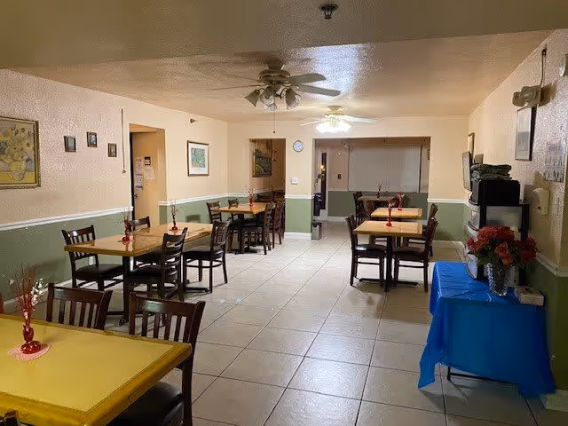 A dining room in Sunny Days Assisted Living with several wooden tables and chairs arranged neatly. Each table has a small red vase with decorative branches. The walls are painted light beige with a green lower half and decorated with framed pictures. There is a ceiling fan with lights, a clock on the wall, and a blue-covered table with flowers and a microwave on the right side.
