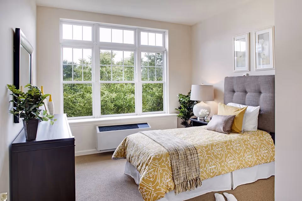 Bright furnished bedroom with a large multi-pane window, upholstered headboard, patterned yellow bedspread, nightstand and dresser.