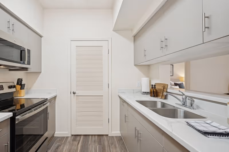 Modern kitchen with gray cabinets, a stainless steel stove and microwave on the left, a double sink with a chrome faucet on the right, and a closed white louvered door at the end. The countertop is white with a marble pattern, and there are kitchen utensils and soap dispensers near the sink. The floor has wood-like vinyl planks.