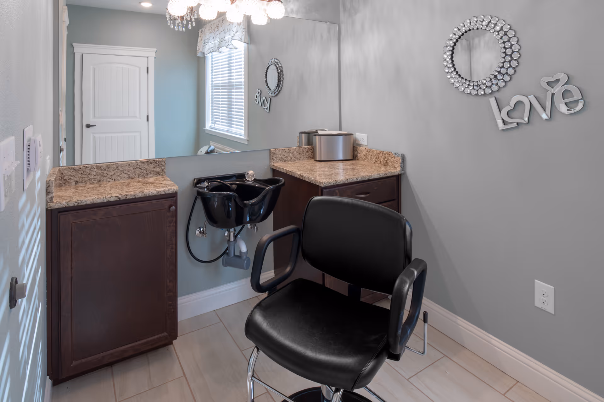 Interior view of a small salon area with a black salon chair in front of a black hair washing sink. The room has light gray walls, a large mirror, a window with blinds and a valance, dark wood cabinets with granite countertops, and a decorative wall mirror with the word 'Love' beside it.