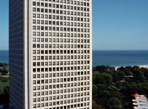 Tall high-rise building facade with a grid of windows overlooking trees and a lake/beach in the background.