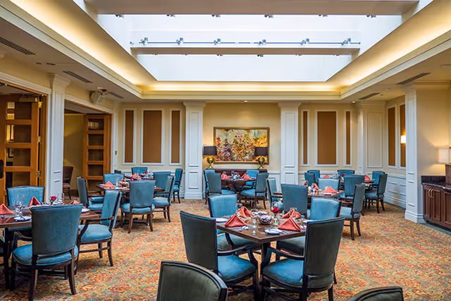 A spacious dining room with multiple round tables set with red napkins, glassware, and silverware. The room features teal upholstered chairs, a patterned carpet, cream-colored walls with decorative paneling, and a large skylight overhead. A colorful painting and lamps are visible on the far wall.