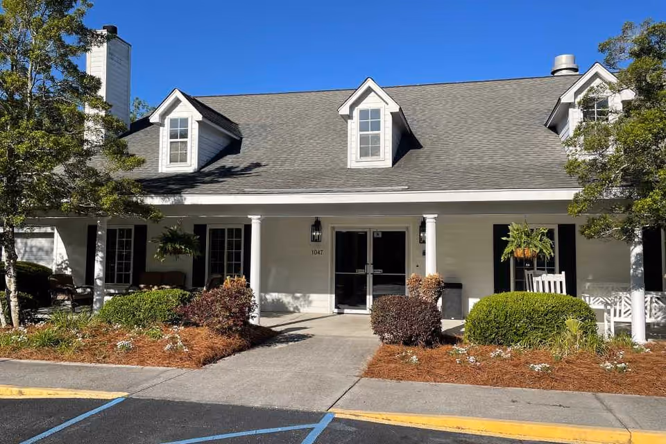 Front exterior view of a single-story building with a gray shingled roof, white walls, and black window shutters. The entrance has double glass doors flanked by white columns and outdoor seating areas with chairs and hanging plants. There are landscaped bushes and trees in front of the building under a clear blue sky.
