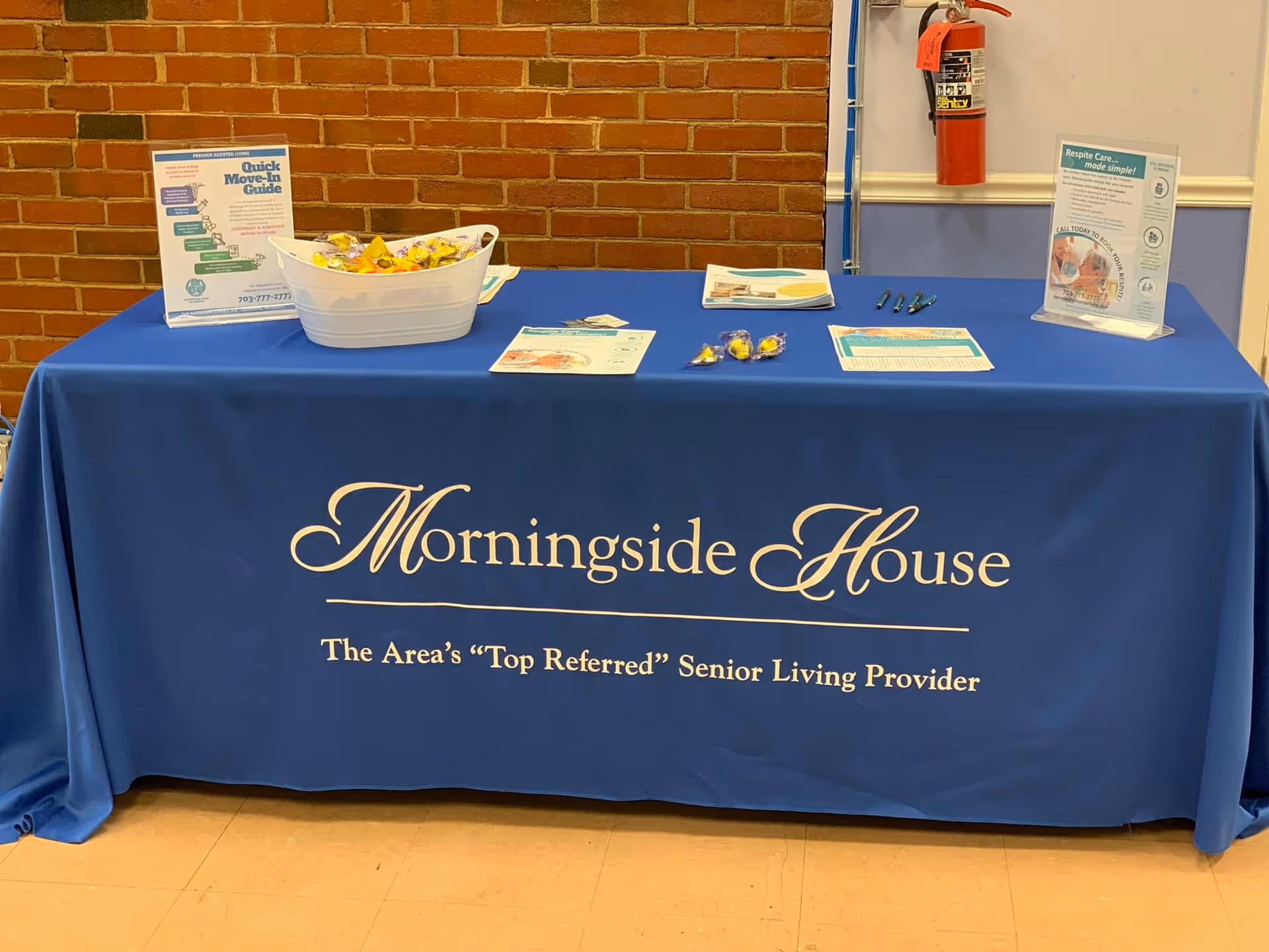 A blue promotional table with a tablecloth reading "Morningside House" displaying brochures, candy, and signage against a brick interior wall.