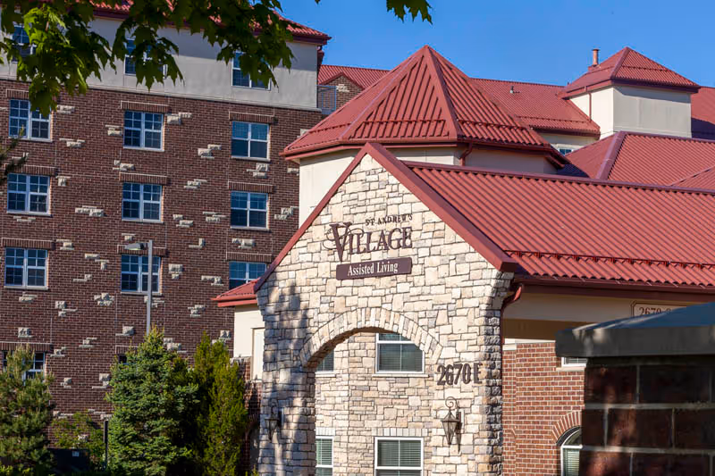 Front exterior of St. Andrew's Village assisted living building with a stone archway and red metal roofs.