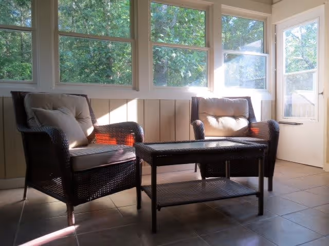 A sunlit room with two cushioned wicker armchairs and a matching wicker coffee table placed on a tiled floor. Large windows let in natural light and show green trees outside. A door with a window is visible on the right side.