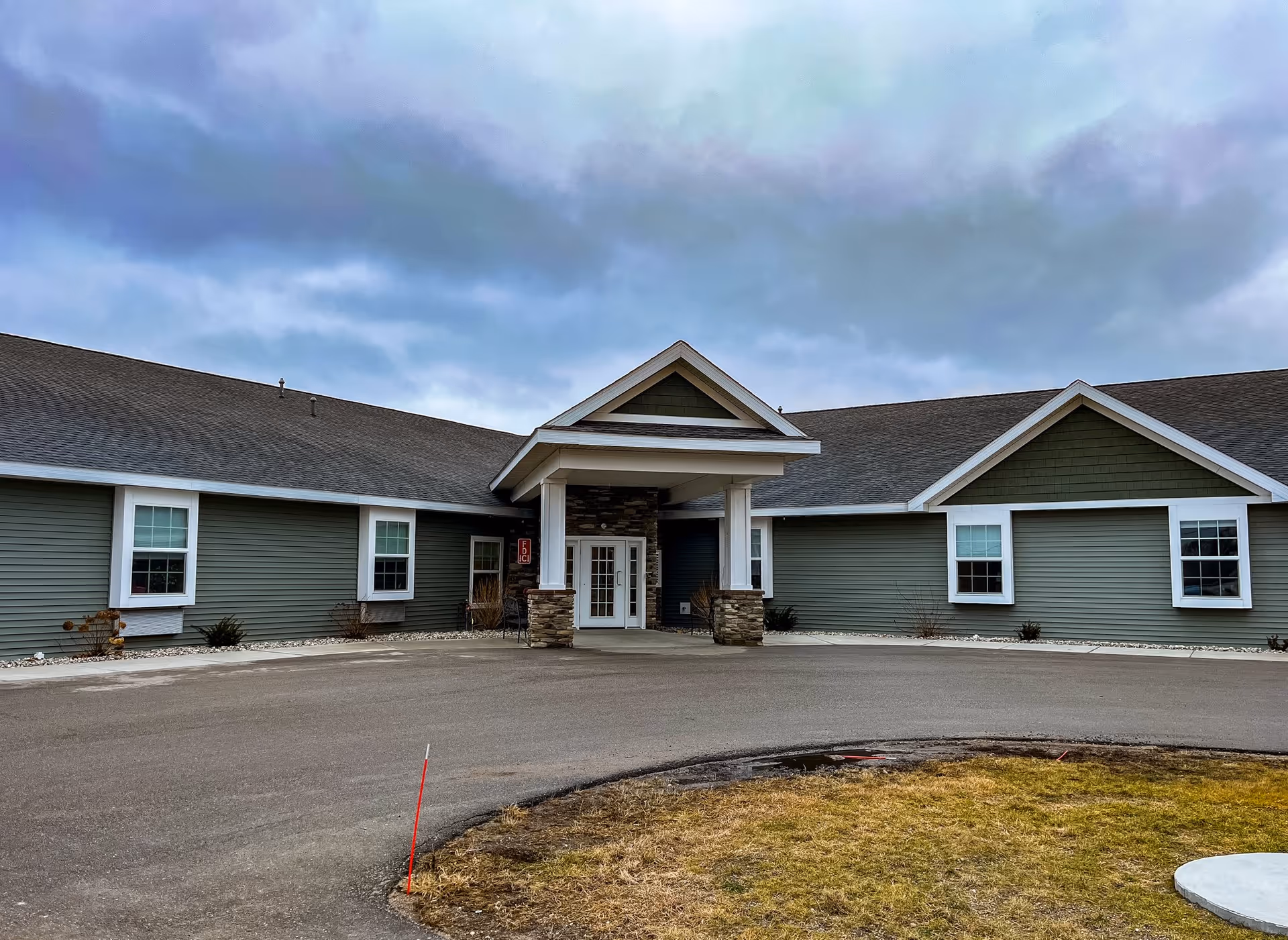 Exterior view of a single-story assisted living facility building with green siding, white trim, and a covered entrance supported by stone pillars. The sky is cloudy and there is a paved driveway and some grass in front.