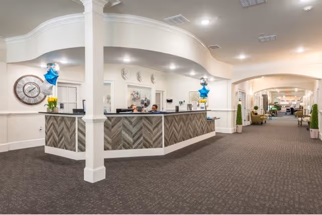 Reception area inside The Mansions at Gwinnett Park Senior Independent Living with a large curved front desk, two staff members seated behind it, decorative wall clock, balloons, and a long carpeted hallway with seating and plants.