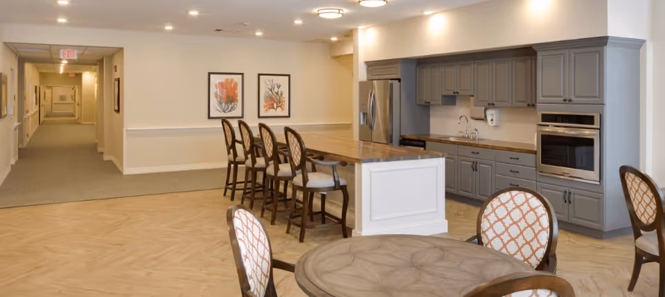 A spacious interior area featuring a modern kitchen with gray cabinets, a stainless steel refrigerator, oven, and a long wooden island with several high chairs. In the foreground, there is a round wooden table with patterned cushioned chairs. The background shows a hallway with carpeted flooring and framed artwork on the walls.