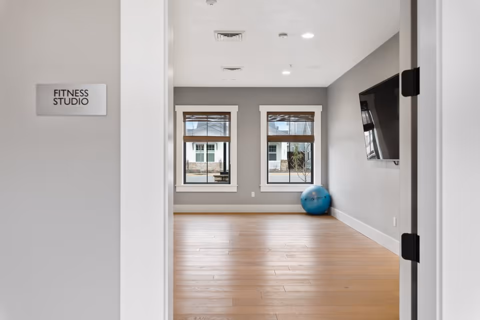 View through an open doorway into a fitness studio with light wood flooring, two windows with brown blinds, a blue exercise ball in the corner, and a wall-mounted flat screen TV. A sign on the left side of the doorway reads 'FITNESS STUDIO'.