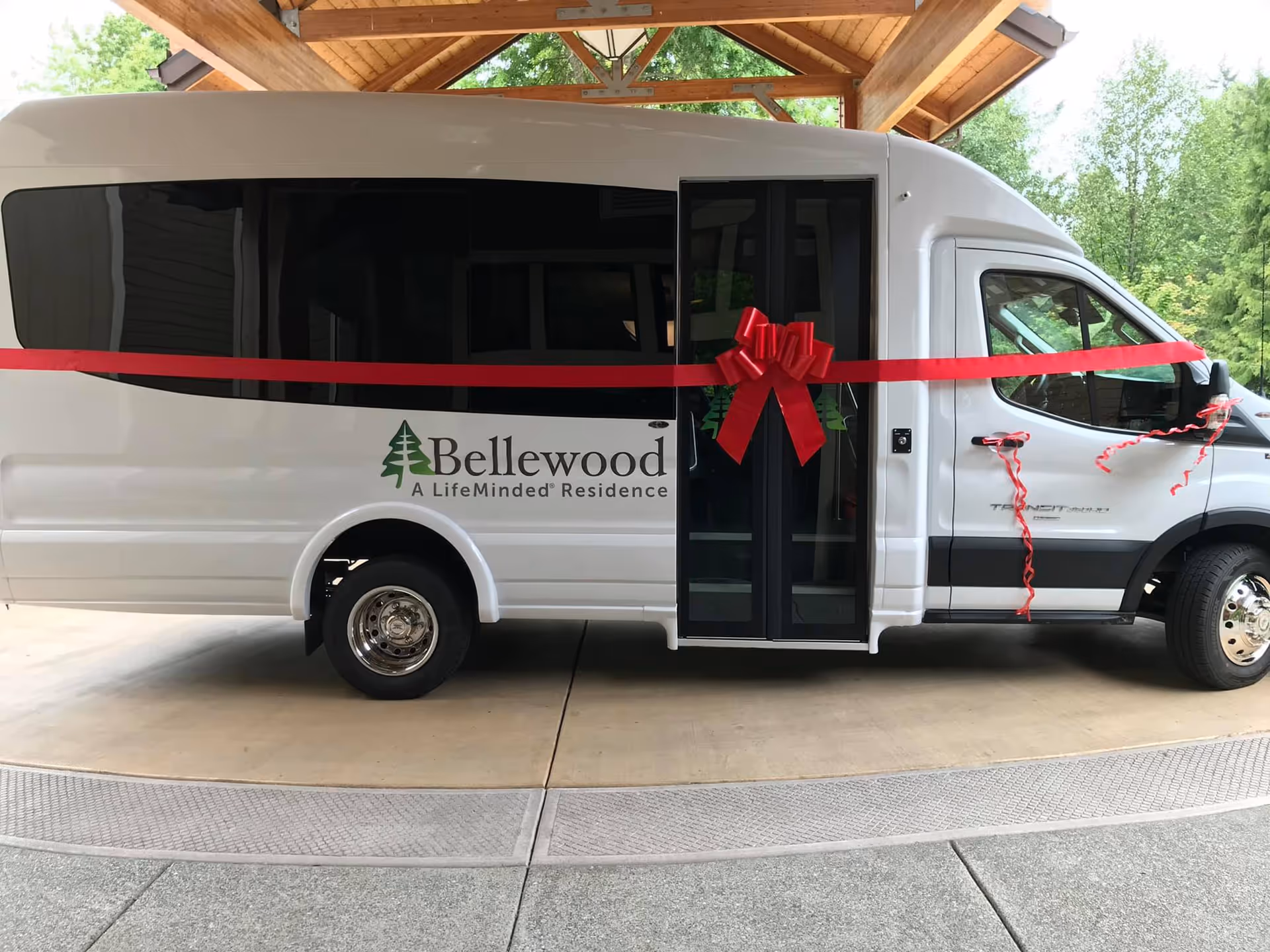 White Bellewood shuttle van decorated with a red ribbon and bow parked under a wooden canopy.