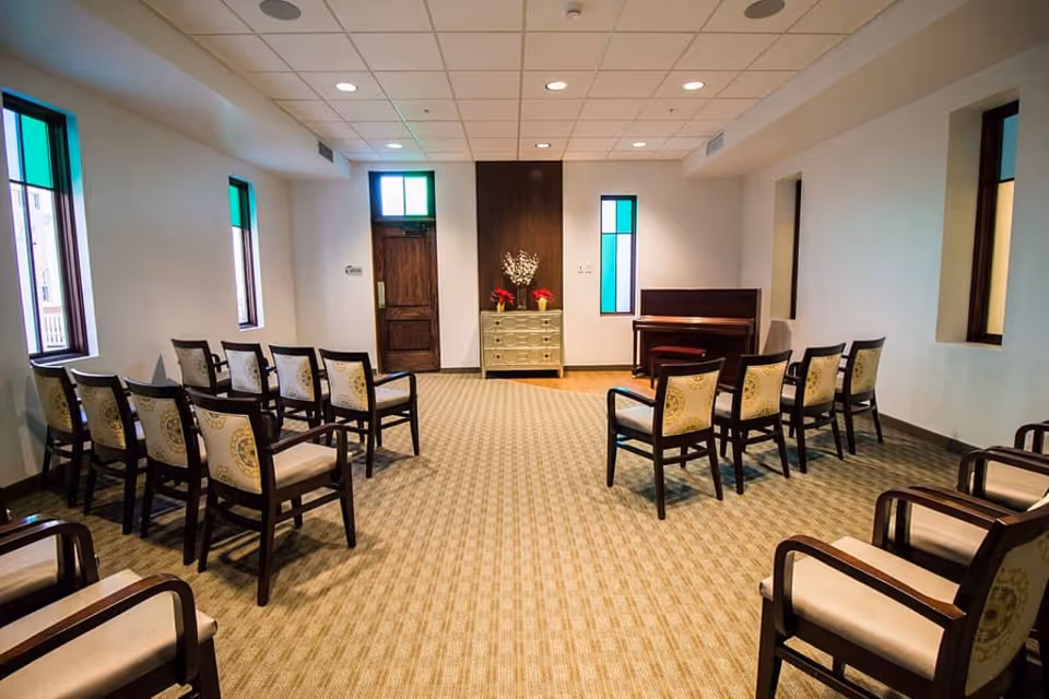 A small meeting or chapel room with rows of wooden chairs arranged facing a small chest of drawers with flower arrangements and a piano against the wall. The room has carpeted flooring, white walls, and several narrow windows with green and blue stained glass panels.