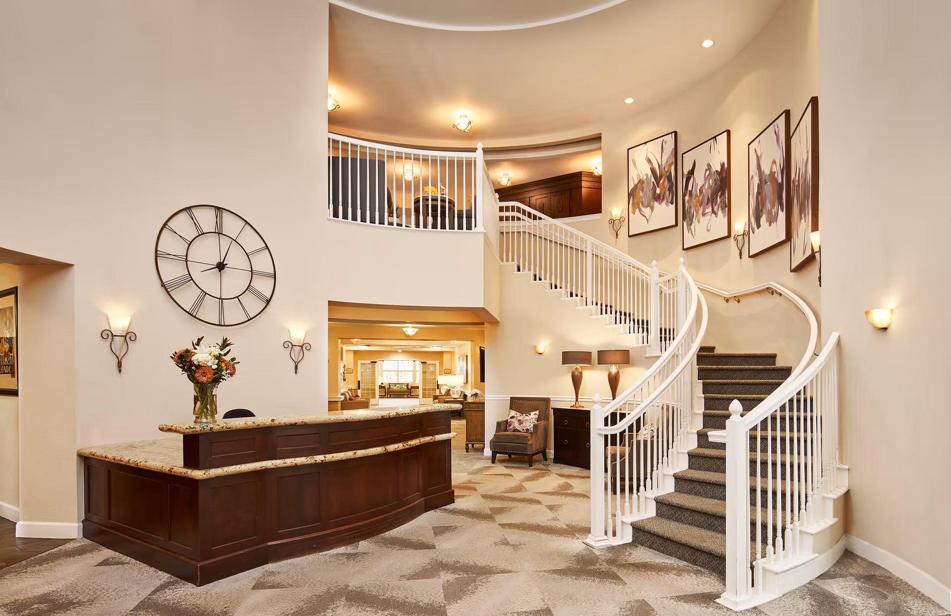 Spacious and well-lit interior lobby area of an assisted living facility featuring a large wooden reception desk with a granite countertop, a decorative wall clock, floral arrangement, a curved staircase with white railings leading to an upper floor, several abstract paintings on the wall, and comfortable seating with a chair and side table with lamps.