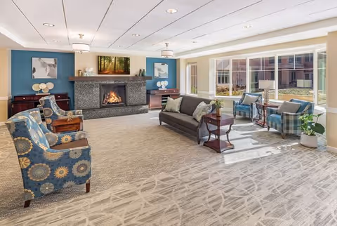 A spacious and well-lit living room in a senior living community featuring a fireplace with a stone surround, a dark gray sofa with cushions, two patterned blue armchairs, two blue plaid armchairs near large windows, side tables with lamps and plants, and artwork on the walls. The room has a beige carpet with a subtle pattern and recessed ceiling lights.